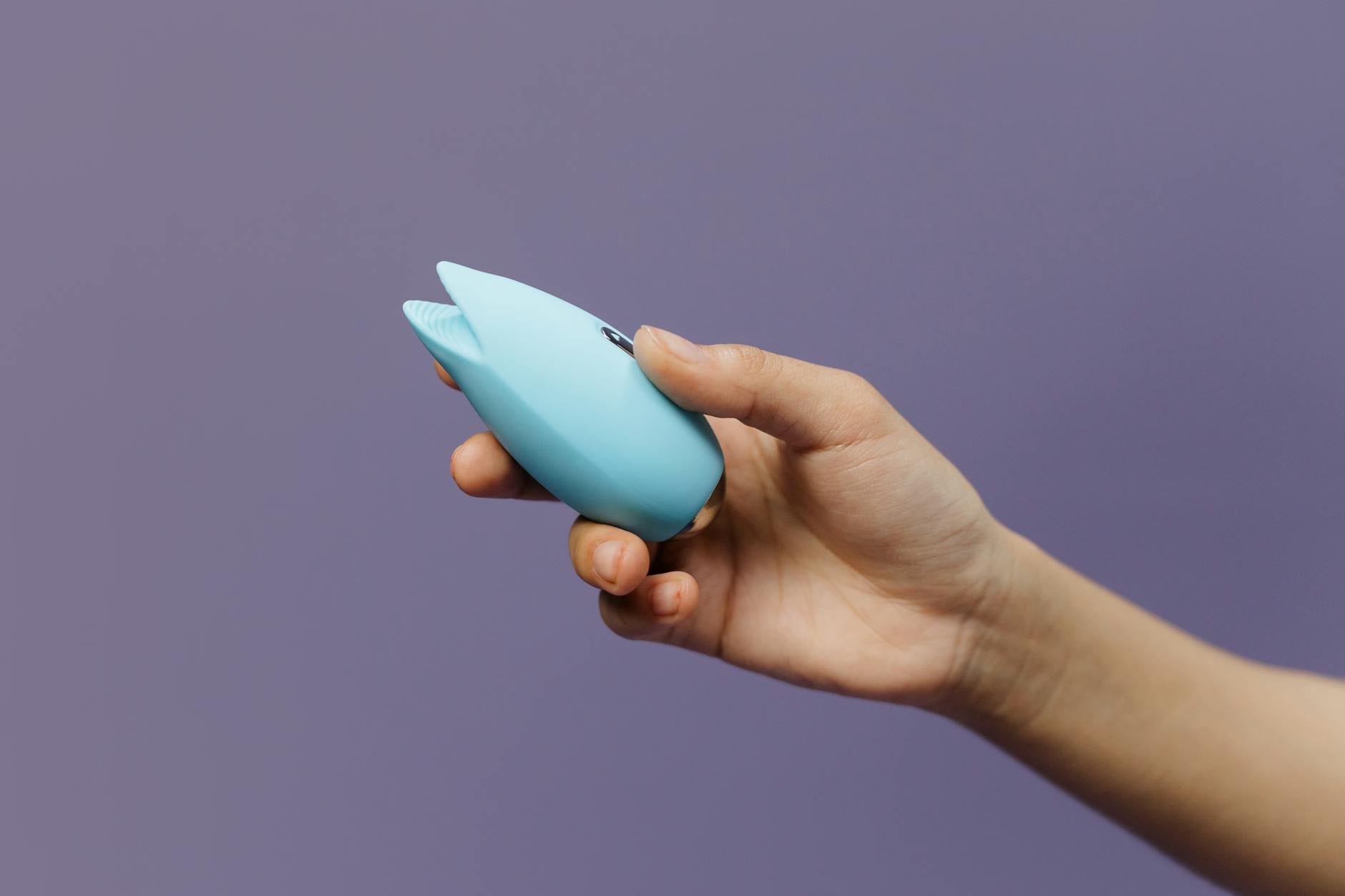 Two hands holding pink and blue silicone vibrators against a pastel background, representing shared pleasure tools.